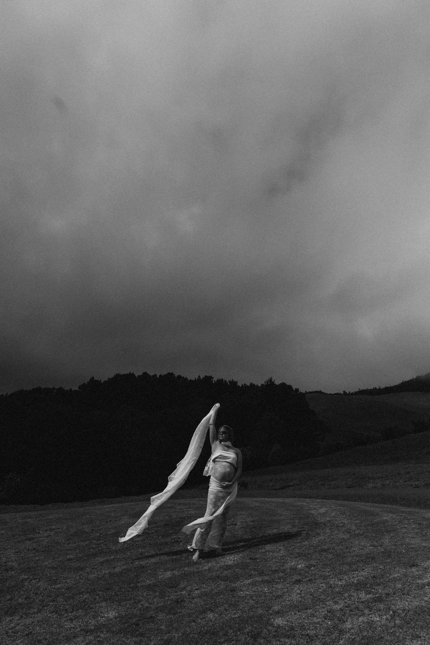 Black and white photo of a pregnant woman in white dress holding flowing fabric against dramatic cloudy sky in open landscape.