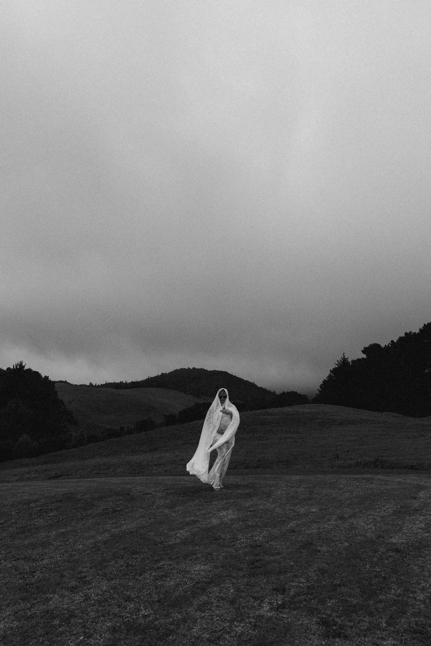 Black and white photo of a person in a white flowing gown standing on a misty hillside landscape.