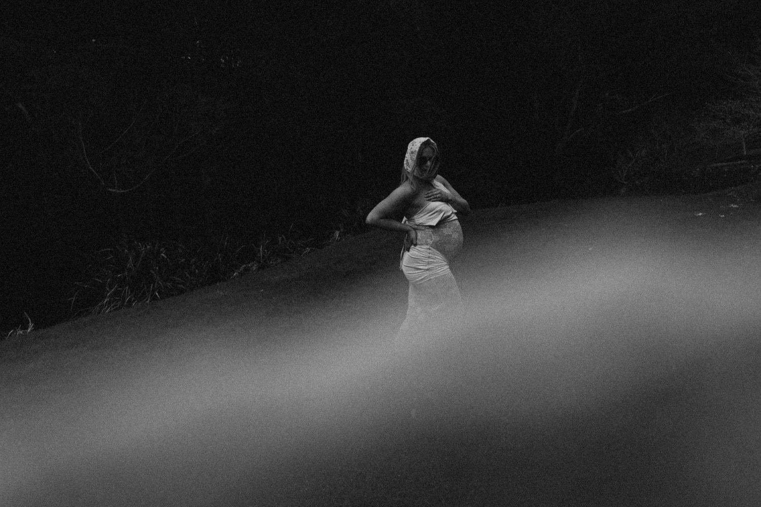 Black and white photo of a pregnant wiman in a white dress standing in a misty, dark forest clearing in Waikato New Zealand.