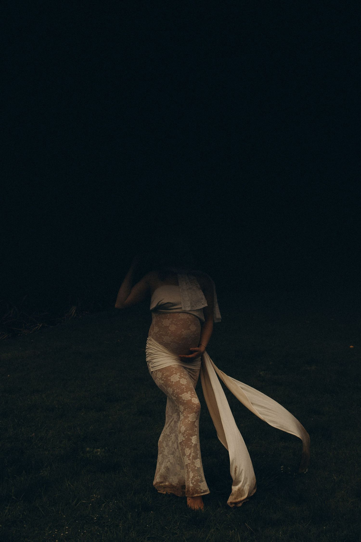Pregnant woman in elegant lace gown standing on dark grass at night, fabric flowing in the breeze.