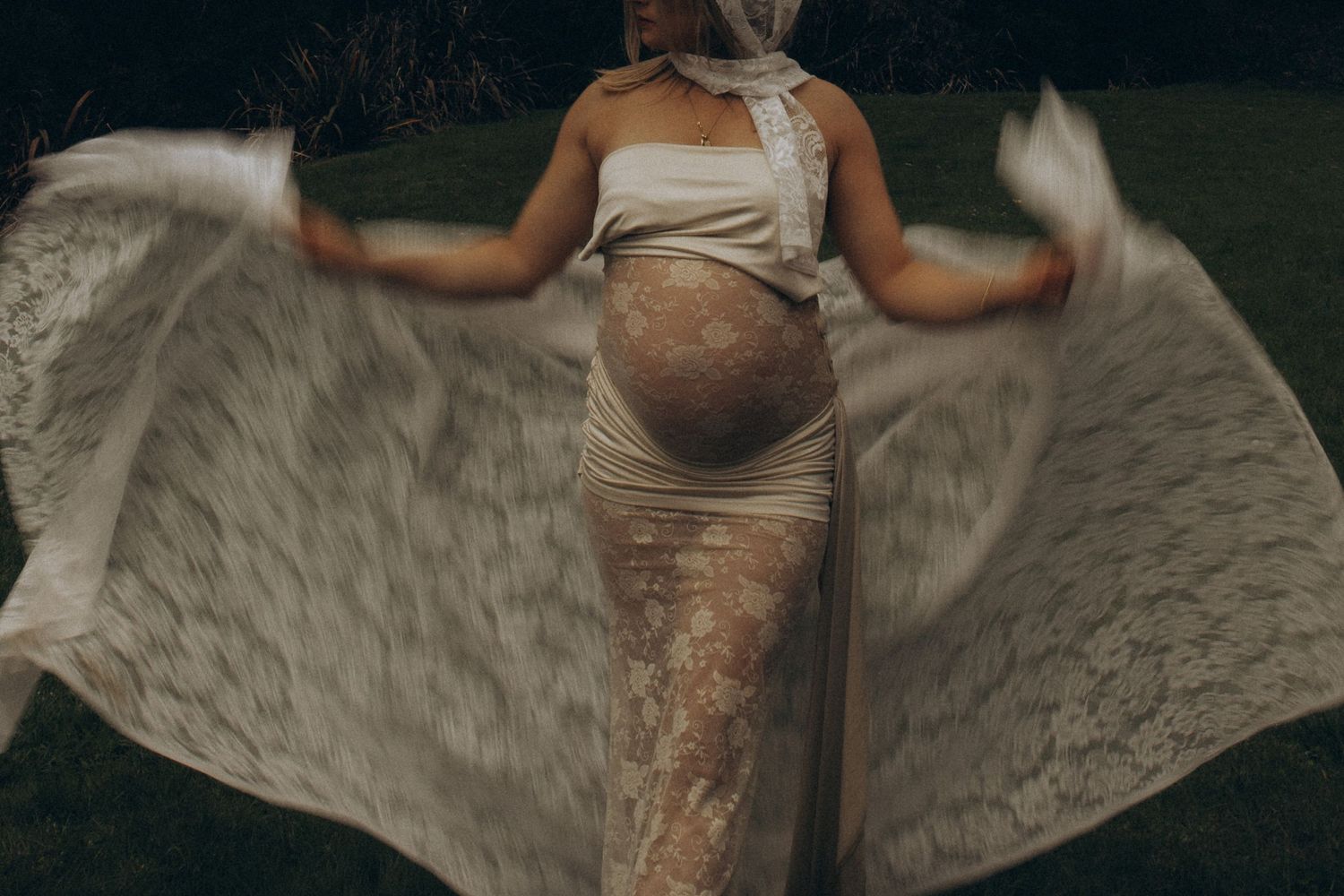 Pregnant woman in lace maternity gown spreading fabric like wings outdoors at dusk in Raglan New Zealand.