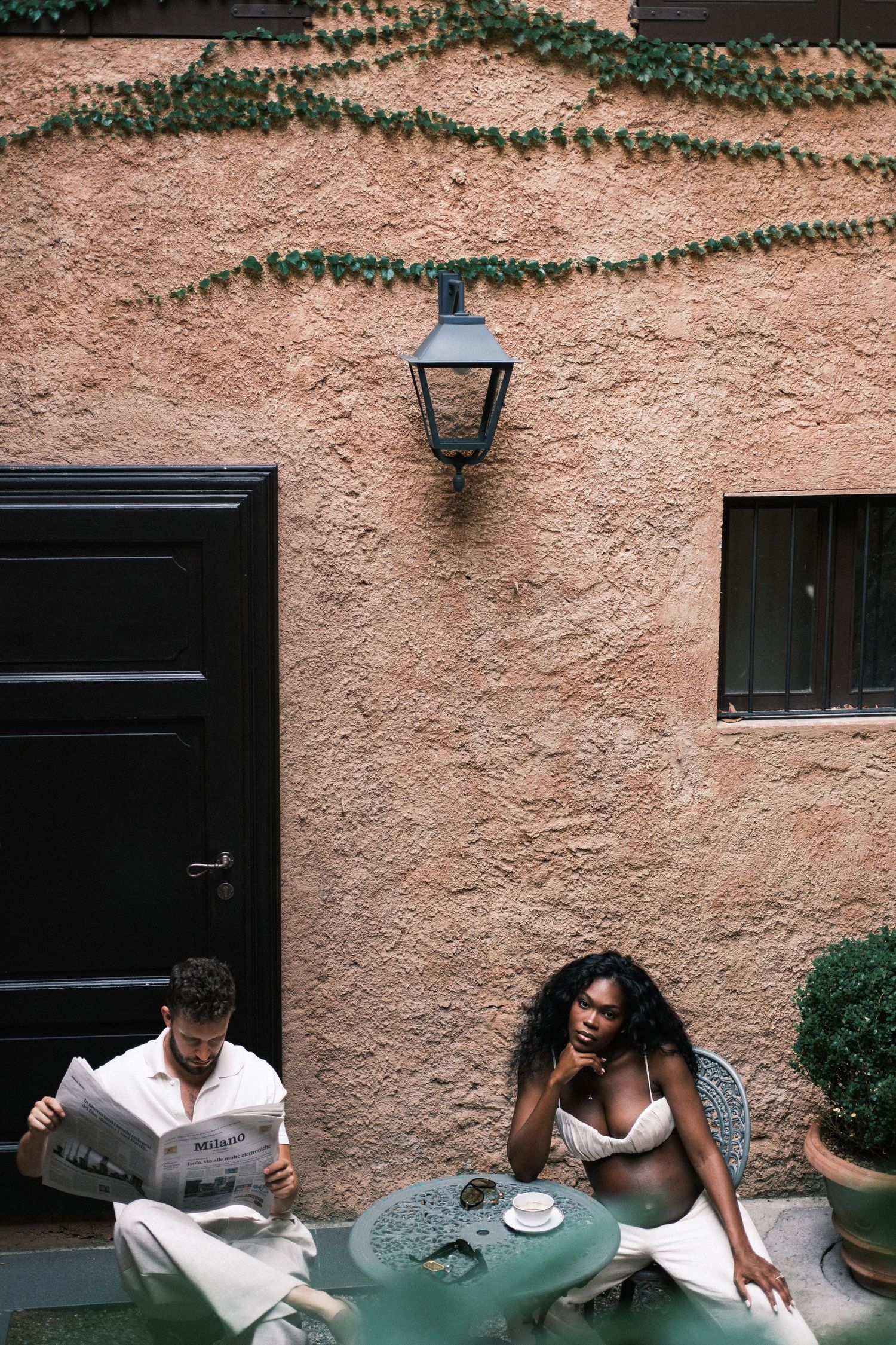 Couple relaxing at outdoor cafe table against textured terracotta wall with ivy, man reading newspaper, woman posing elegantly.