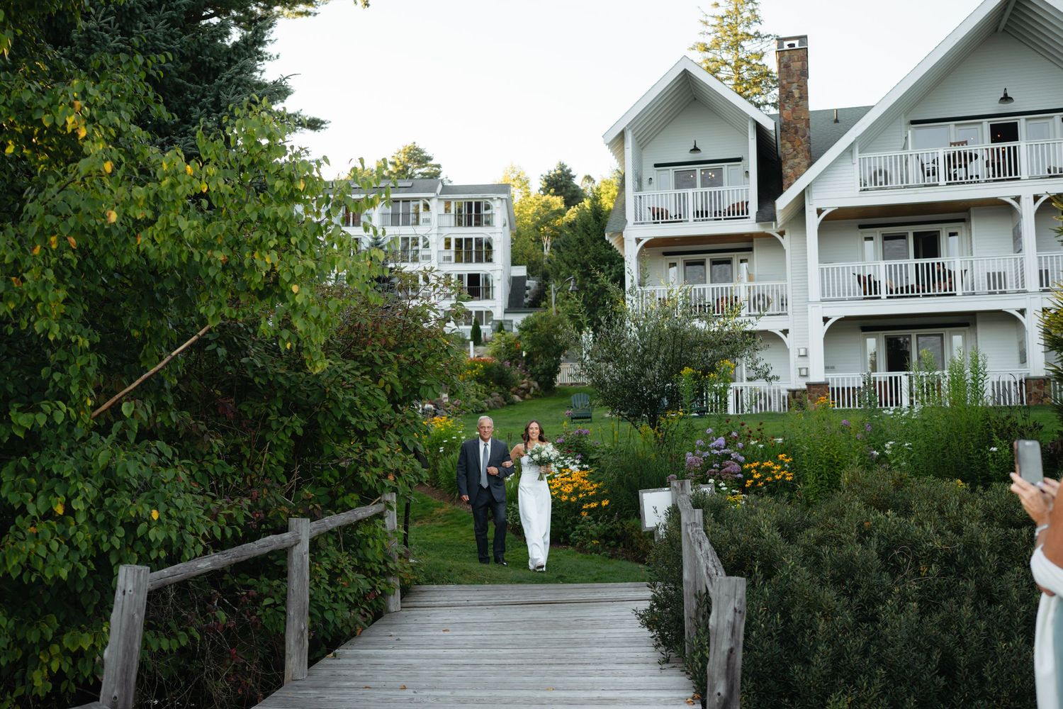Bride and groom walk along wooden bridge path surrounded by lush gardens with elegant white resort building backdrop.