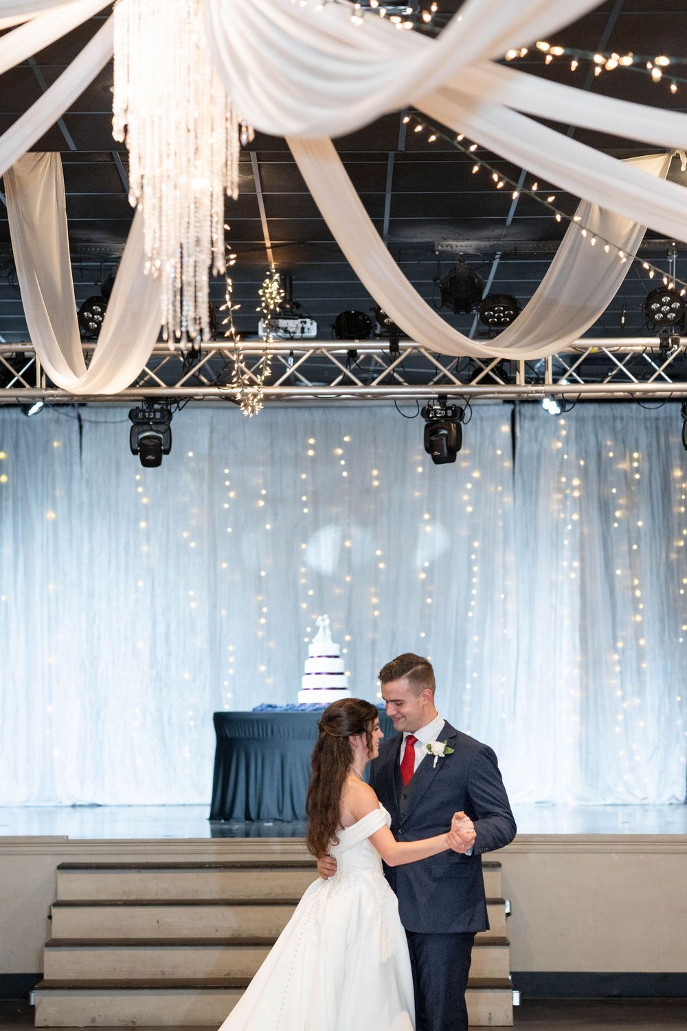 Bride and groom share first dance in elegant wedding venue with white draping, fairy lights, and tiered wedding cake backdrop.