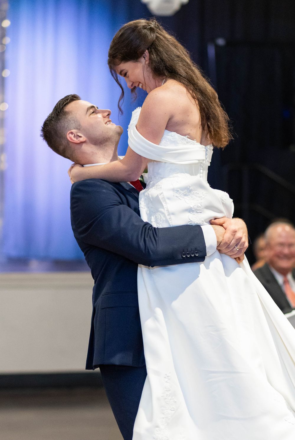 Groom in navy suit lifts bride in white off-shoulder gown during joyful first dance at wedding reception.