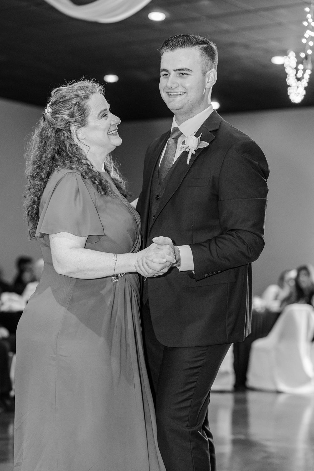 Black and white photo of a groom dancing with his mother at a wedding reception, both smiling joyfully.