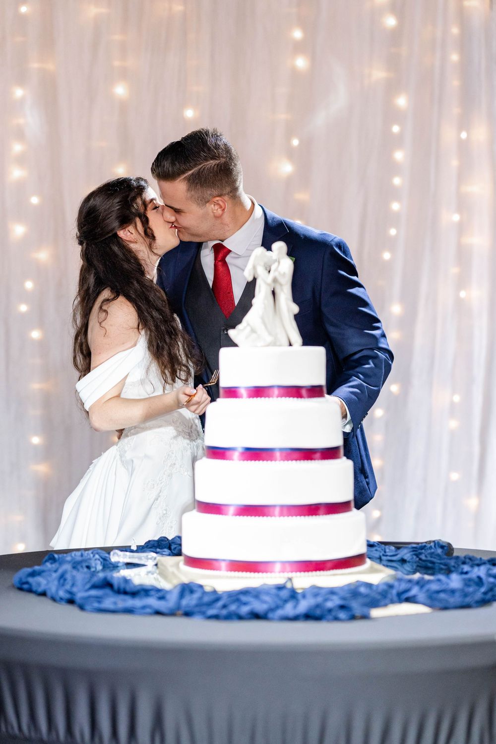 Newlyweds kissing behind a white four-tier wedding cake with red ribbons and couple topper at their reception.