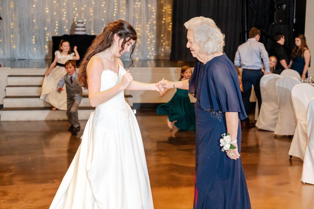 Bride in white gown dancing with elderly woman in navy blue dress at elegant wedding reception.