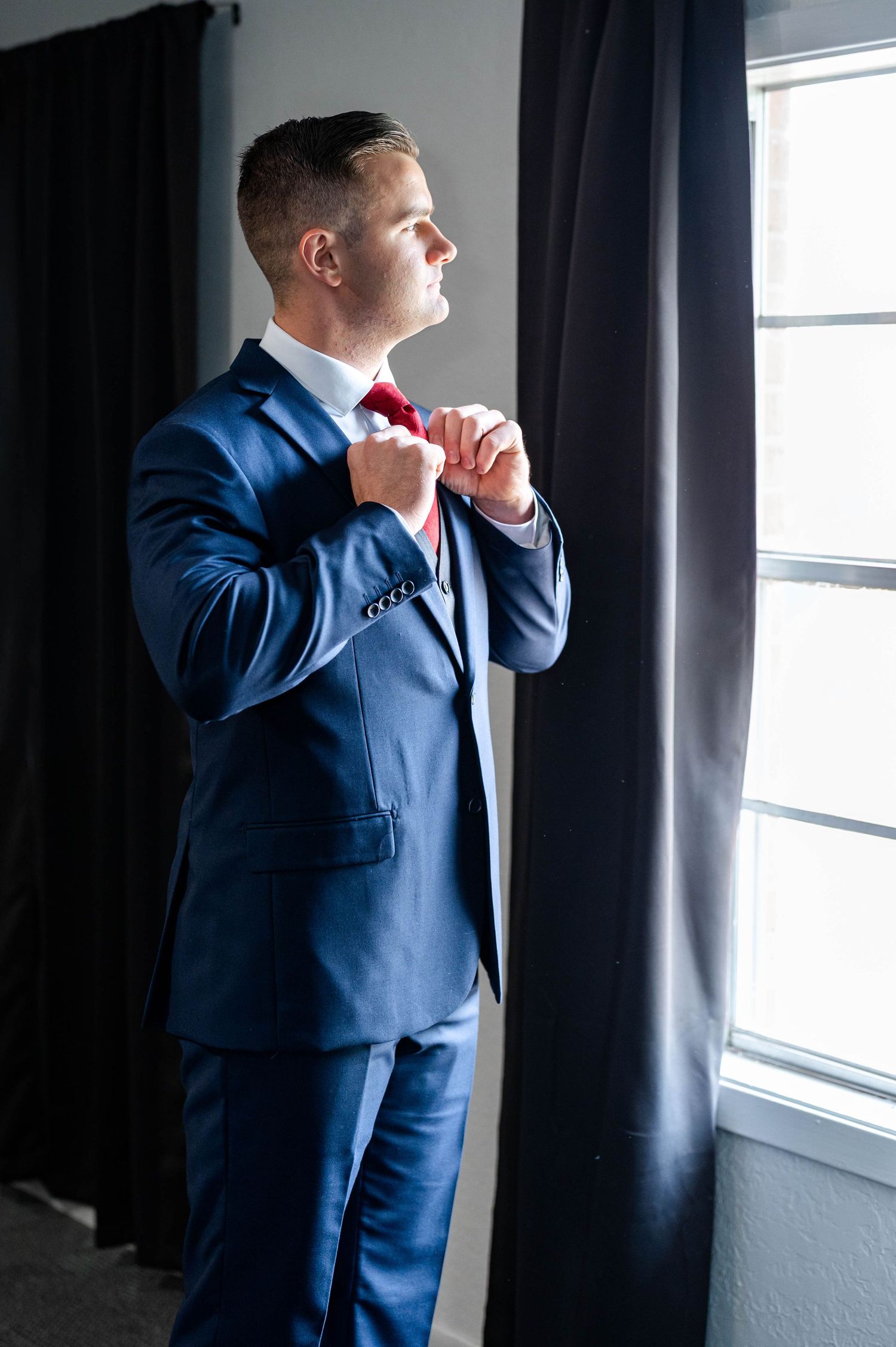 A groom in a navy blue suit adjusts his red bow tie while standing by a window in soft natural light.