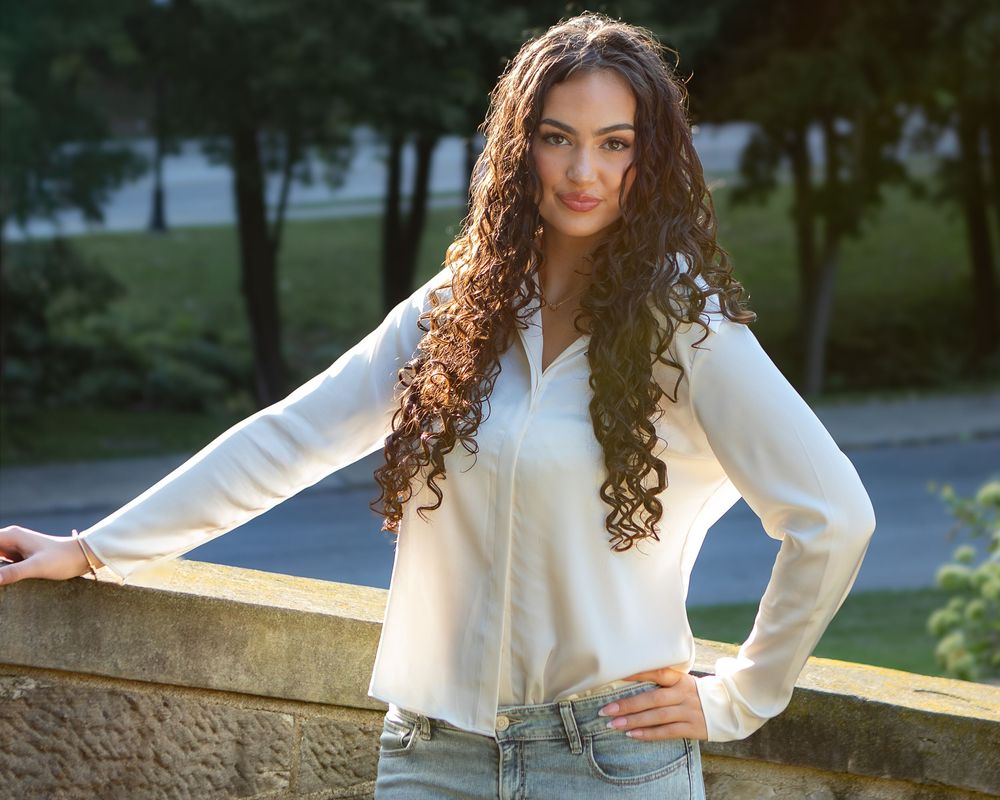 Young woman with long curly brown hair wearing a white blouse and jeans, posing outdoors in a park setting.