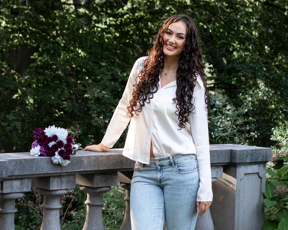 Young woman with curly hair in white blouse and jeans poses by stone railing with burgundy flowers outdoors.