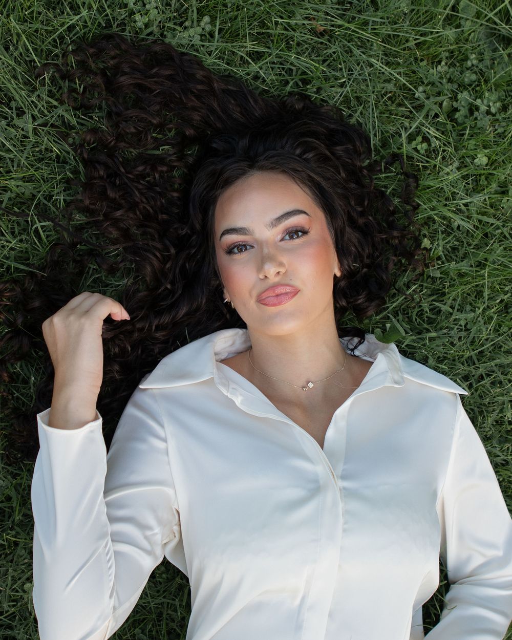 Woman with dark curly hair lying on green grass wearing a white button-up shirt.
