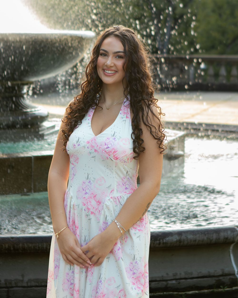 Young woman in white floral halter dress smiling in front of a decorative fountain outdoors.
