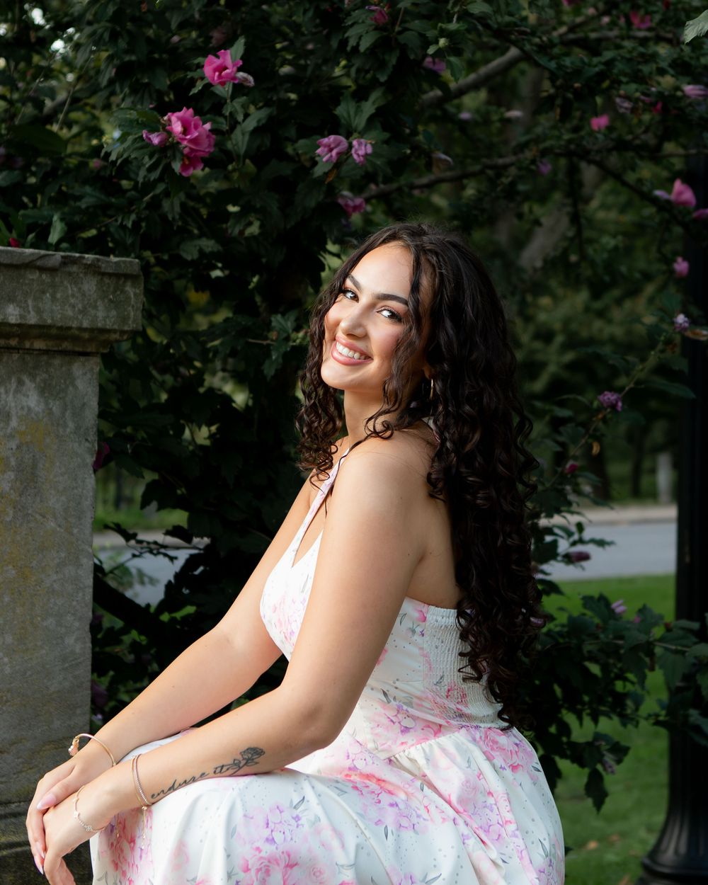 Smiling young woman with curly hair in floral dress sits near blooming pink flowers outdoors.
