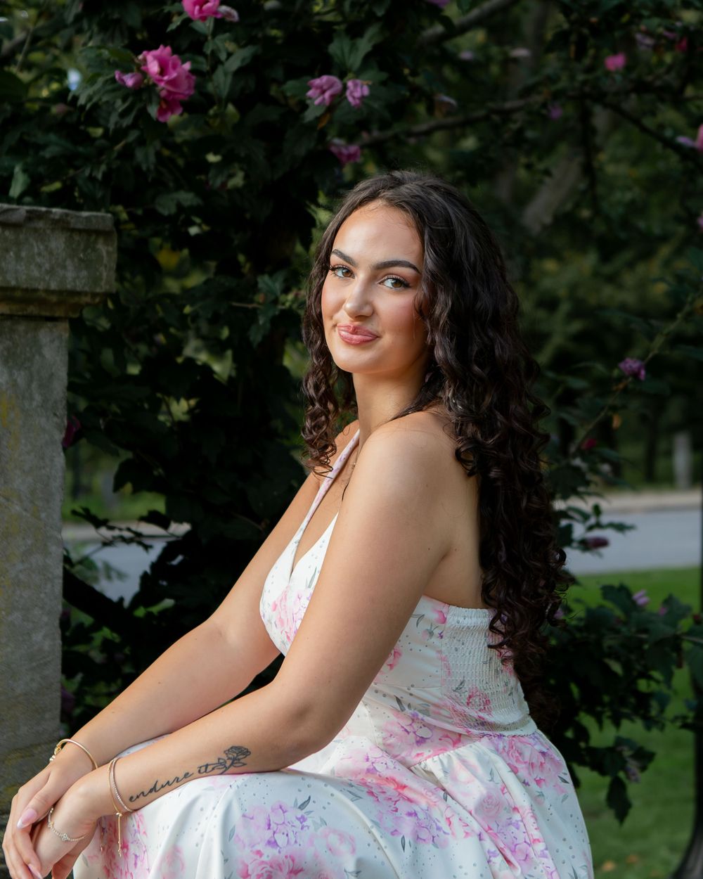 Young woman in floral dress with curly hair sitting outdoors near blooming pink flowers and stone wall.