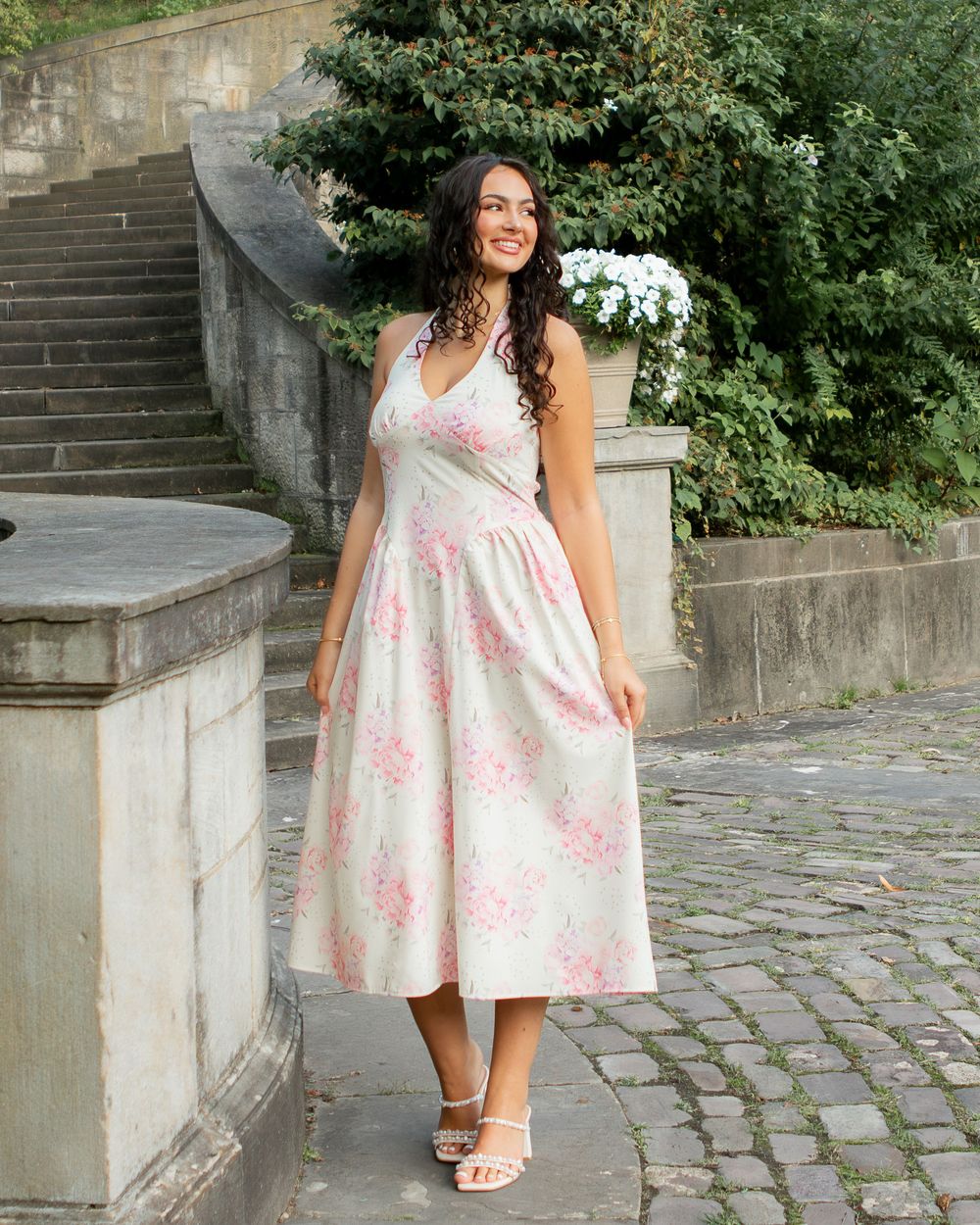 Woman in a floral midi dress standing on cobblestone steps in an outdoor garden setting.