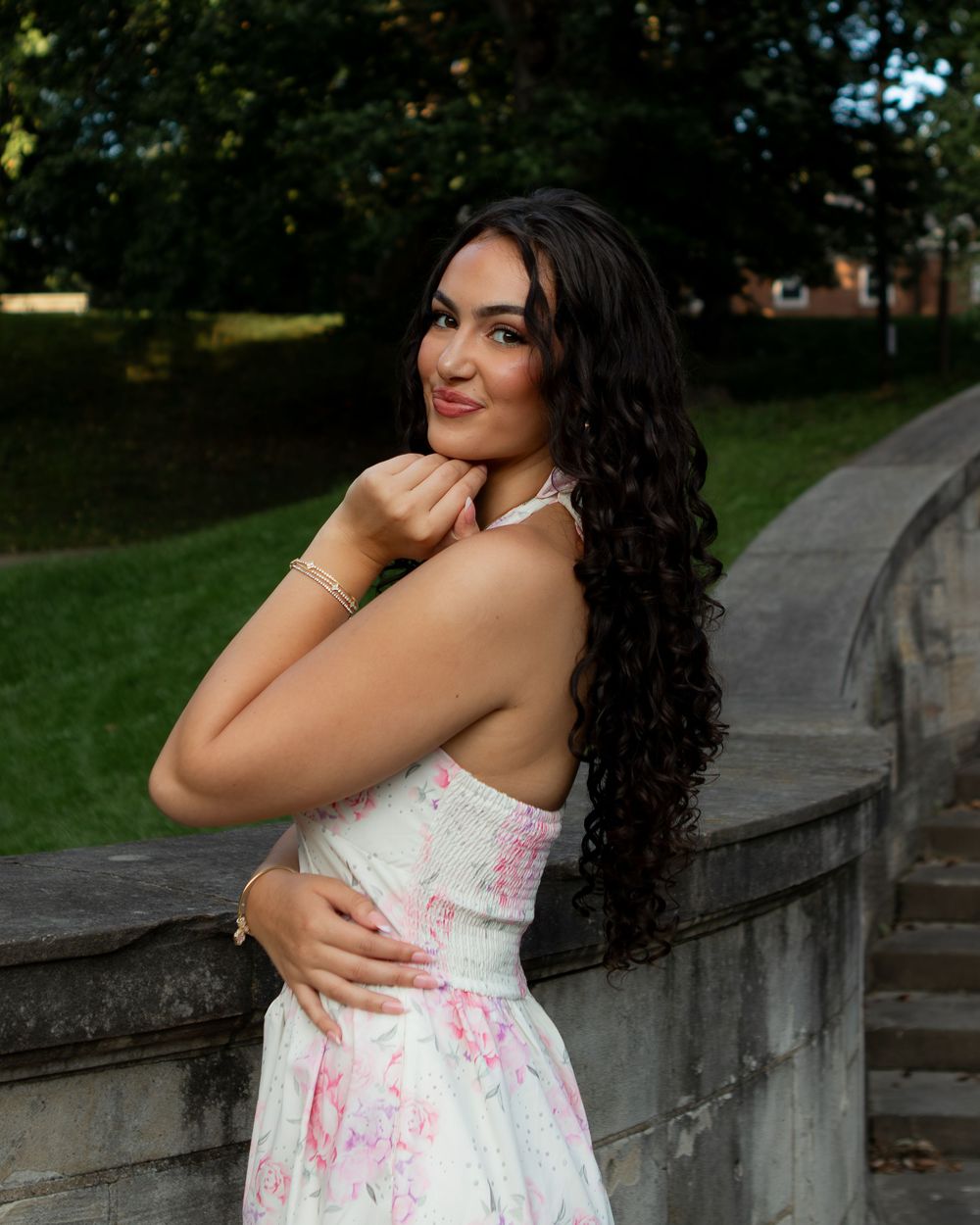 Young woman in floral dress poses gracefully outdoors near stone railing with lush green trees in background.