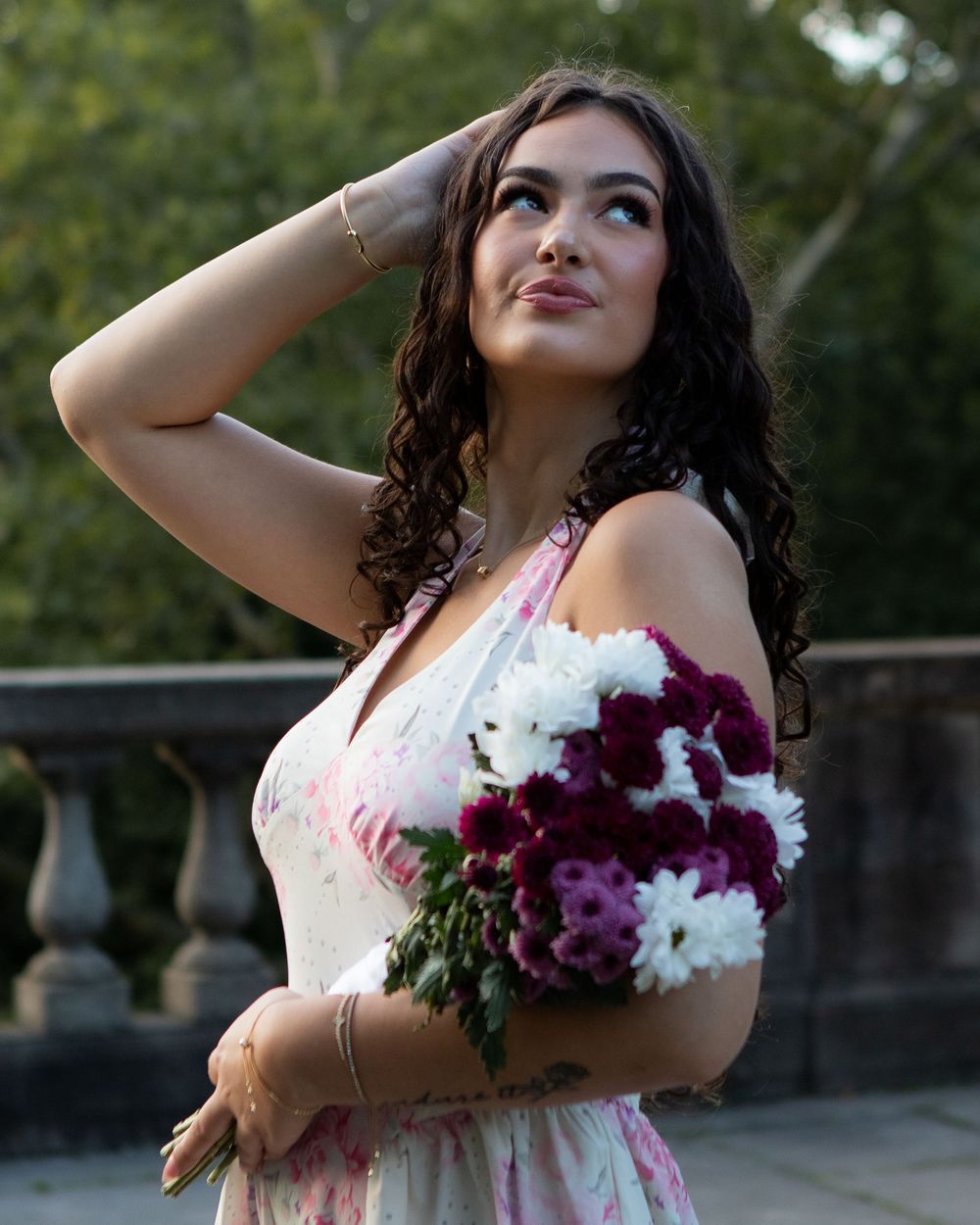 Woman in a floral halter dress holding white and purple flowers while looking upward outdoors.