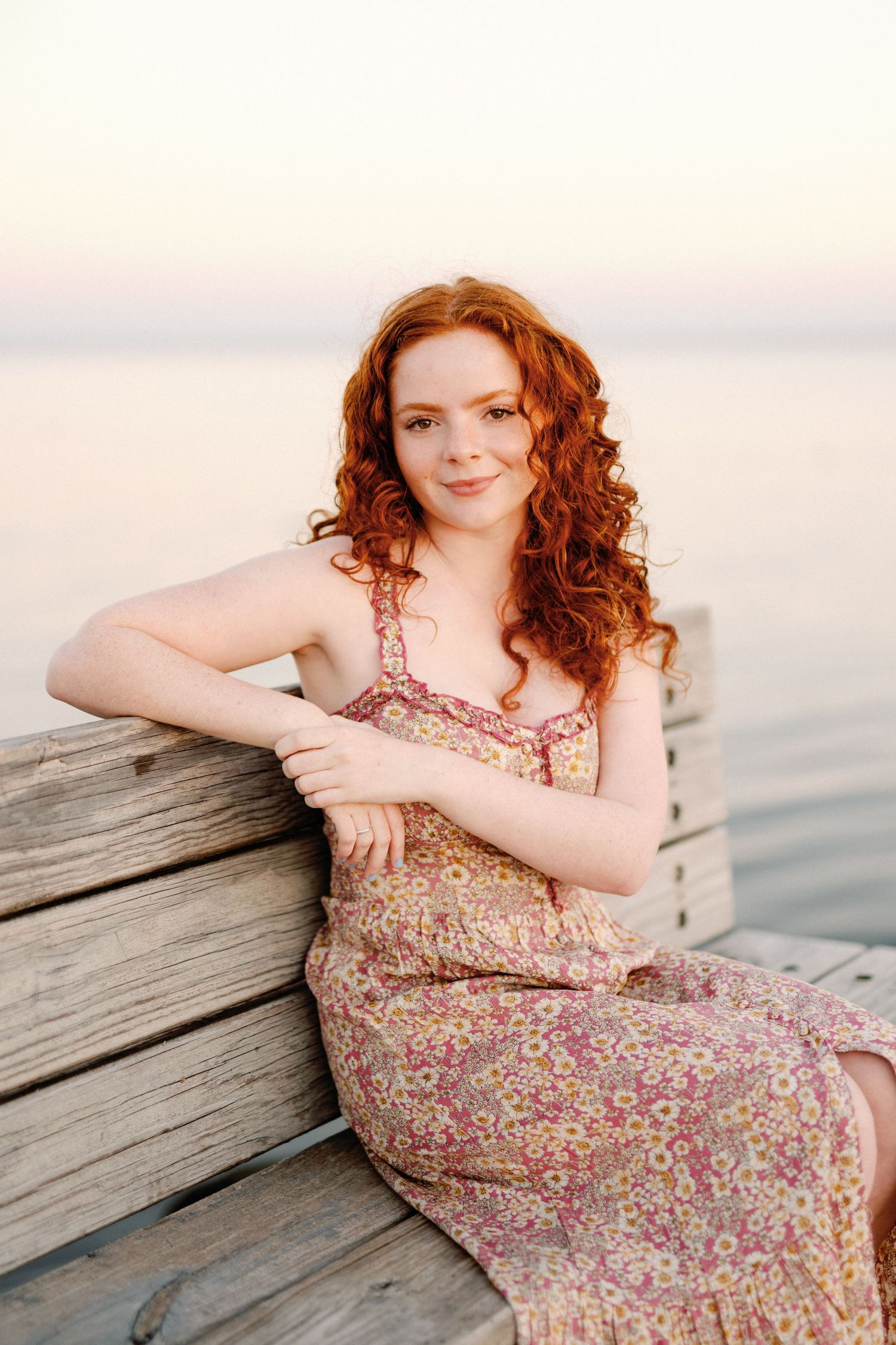 Young woman with curly red hair in floral sundress sitting on wooden dock by calm water at sunset.