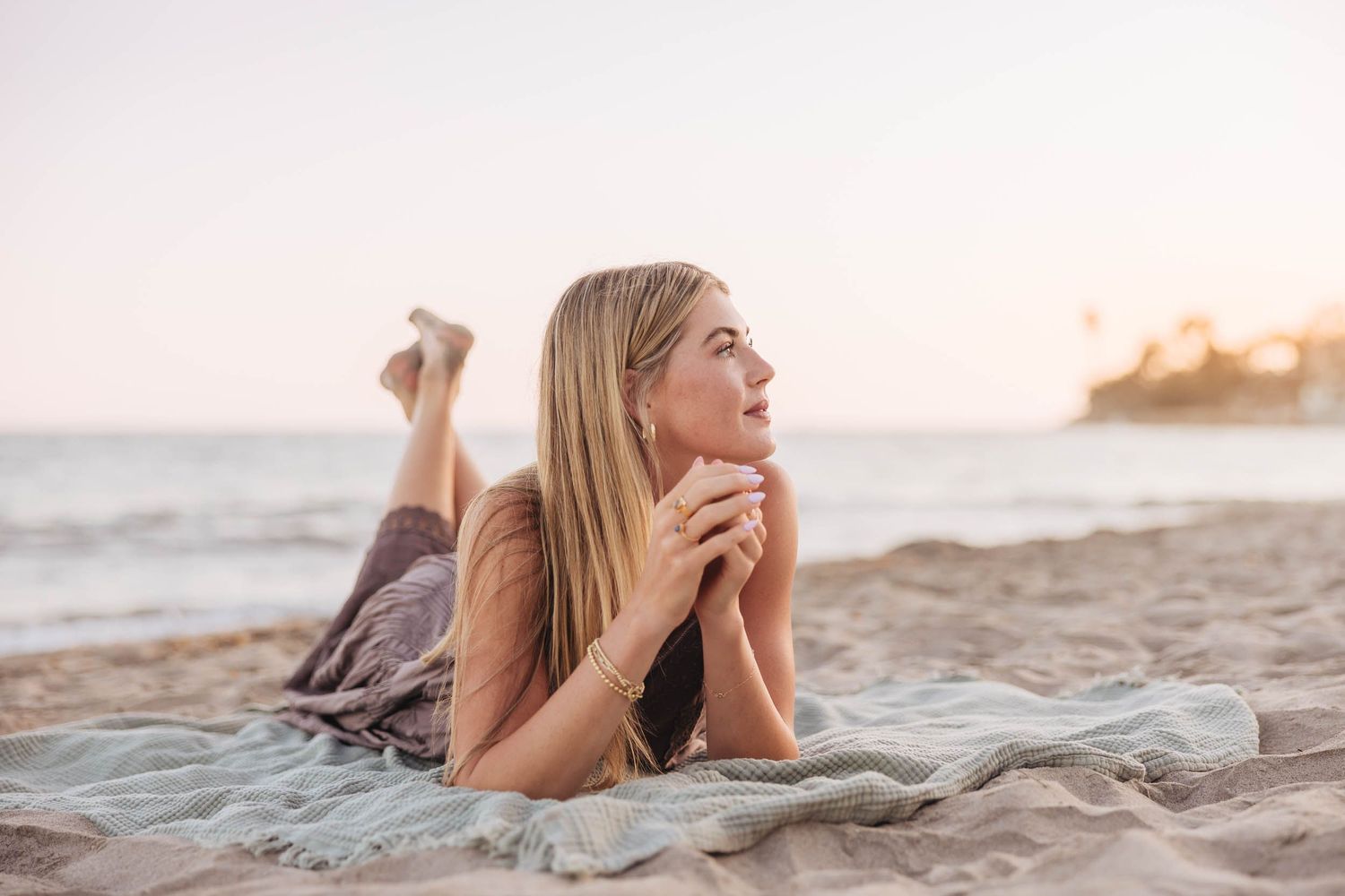 Young blonde woman lying on sandy beach at sunset, gazing thoughtfully at the ocean horizon.