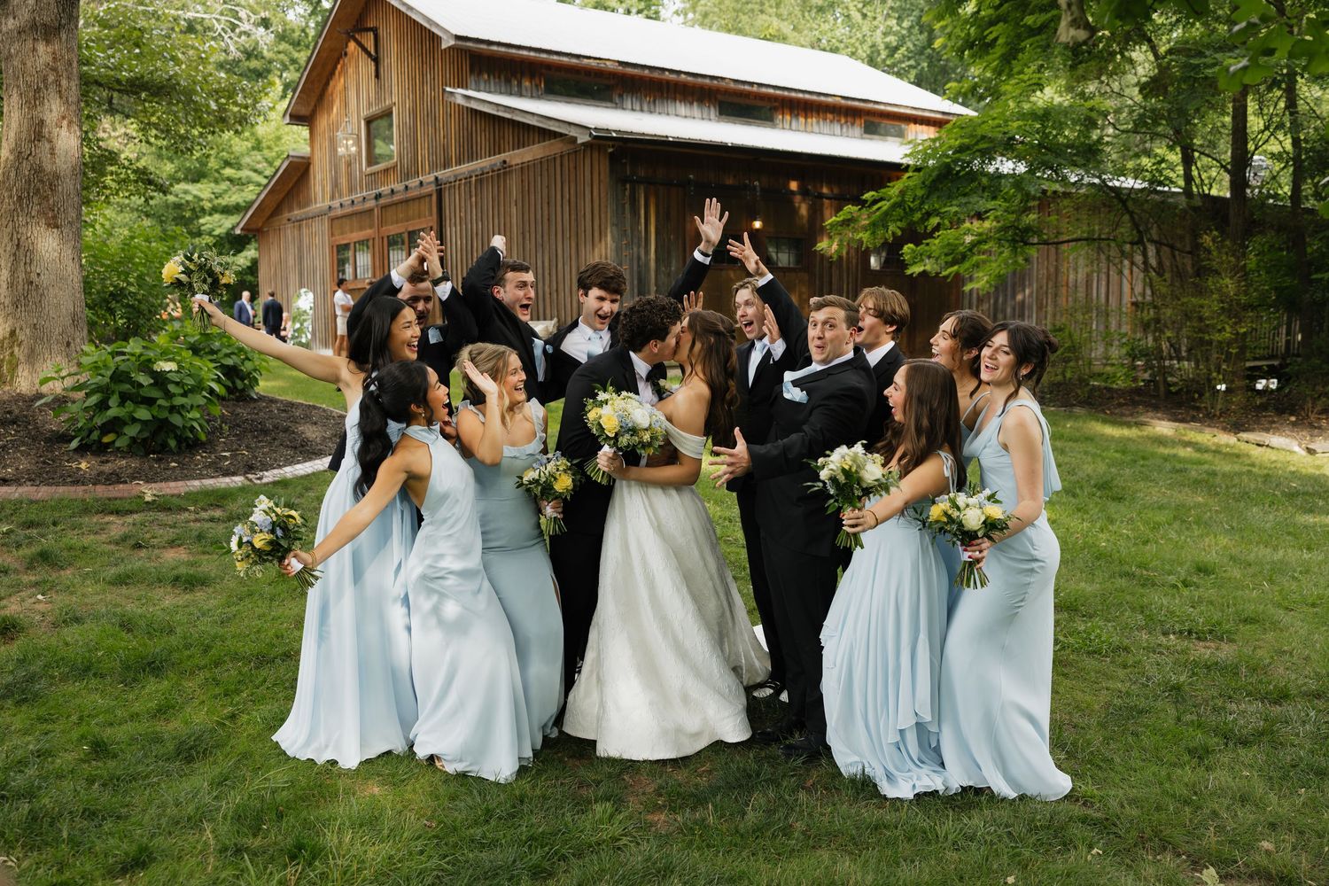 Wedding party celebrating outdoors near rustic barn, bridesmaids in light blue gowns surround couple in joyful group photo.