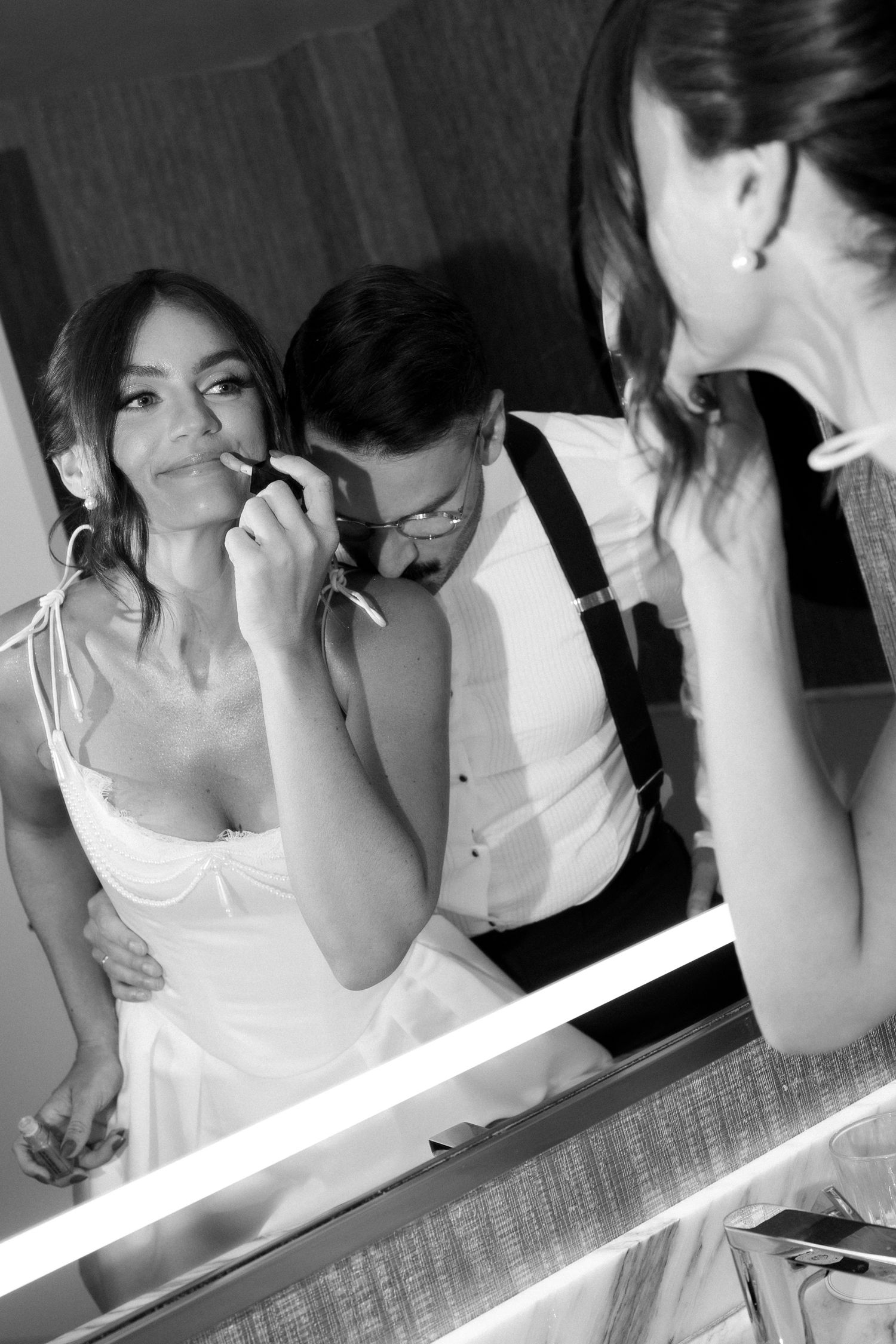 Black and white photo of a bride applying lipstick with groom behind her, reflected in a lit vanity mirror.