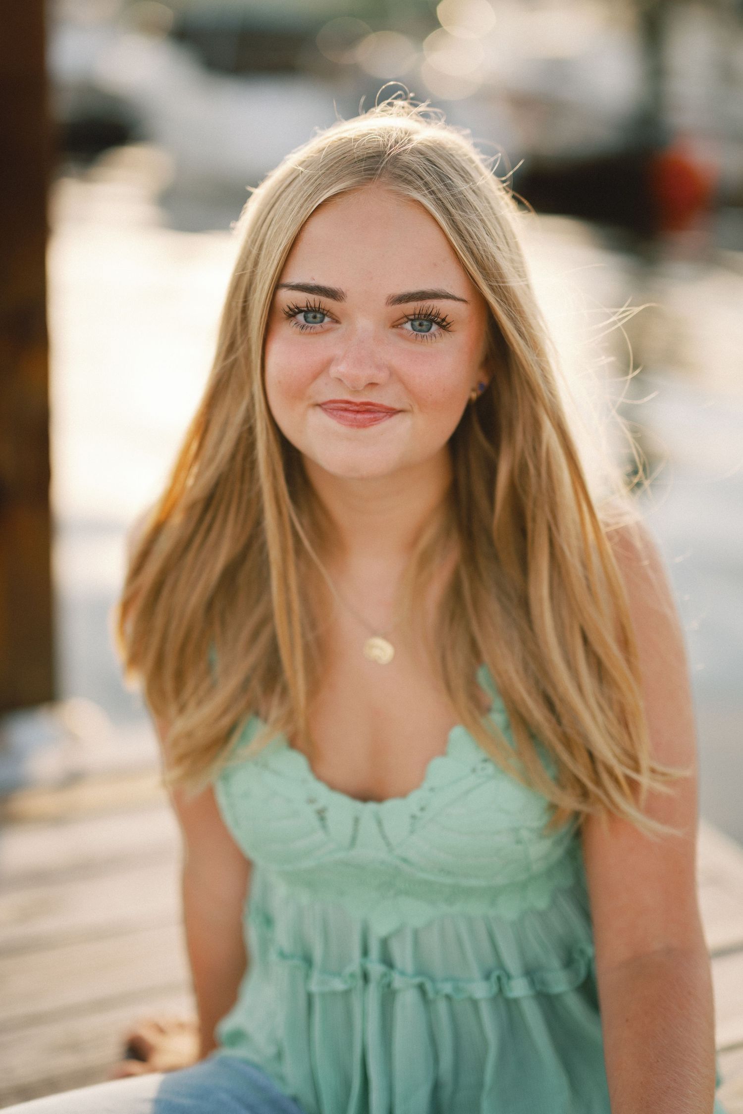Young woman with curly brown hair smiling outdoors at beach, wearing floral top in natural sunlight.