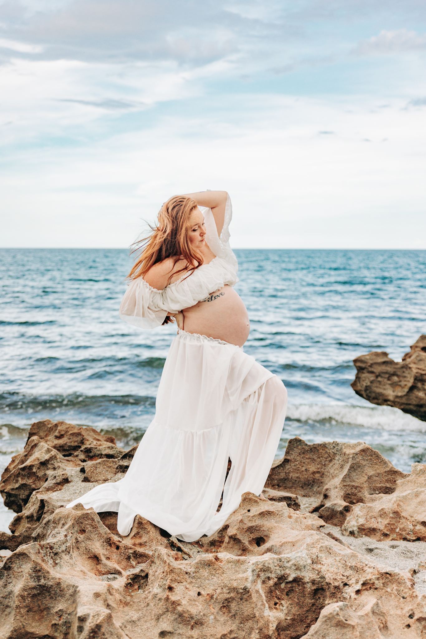 Maternity photographer Jupiter FL capturing an expecting mother in a flowing white dress on the rocky shore at Coral Cove Beach.