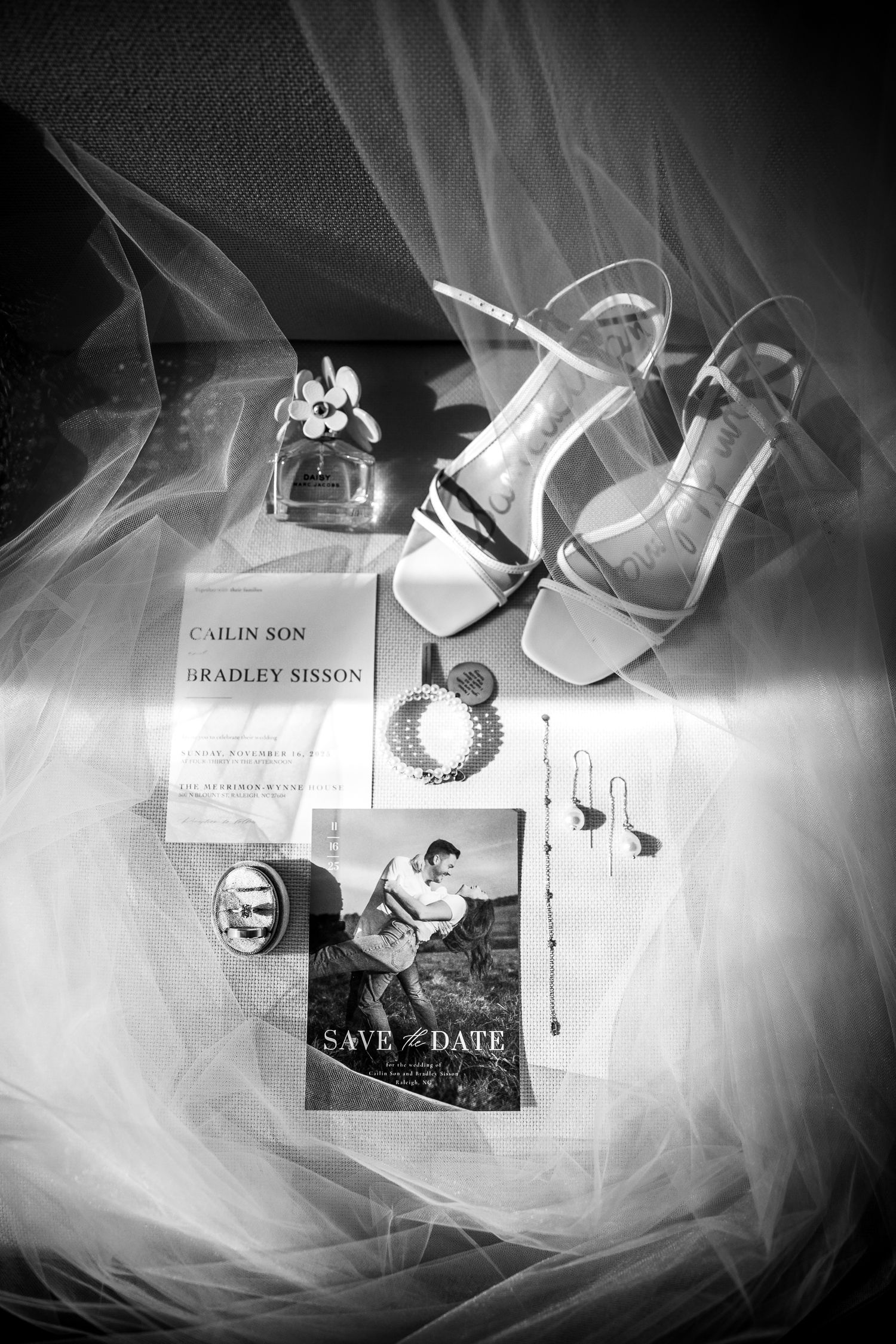 Black and white flat lay of wedding details including save the date cards, bridal shoes, jewelry, and perfume on tulle fabric.