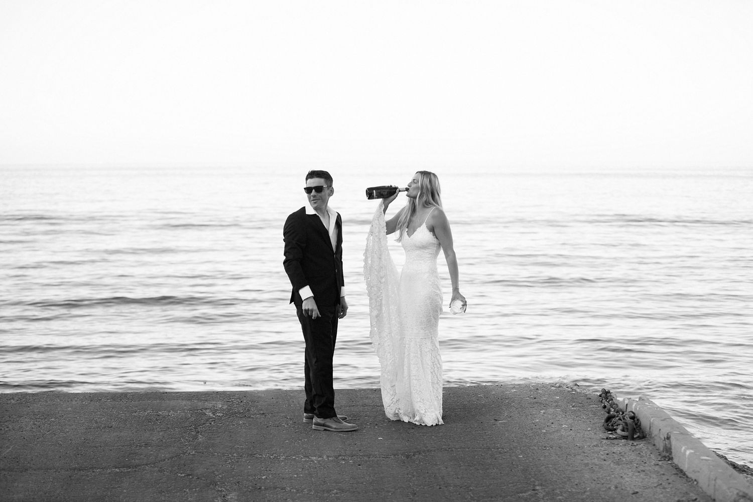 Black and white photo of a couple in formal wear standing by the ocean, woman in white gown drinking from a bottle.