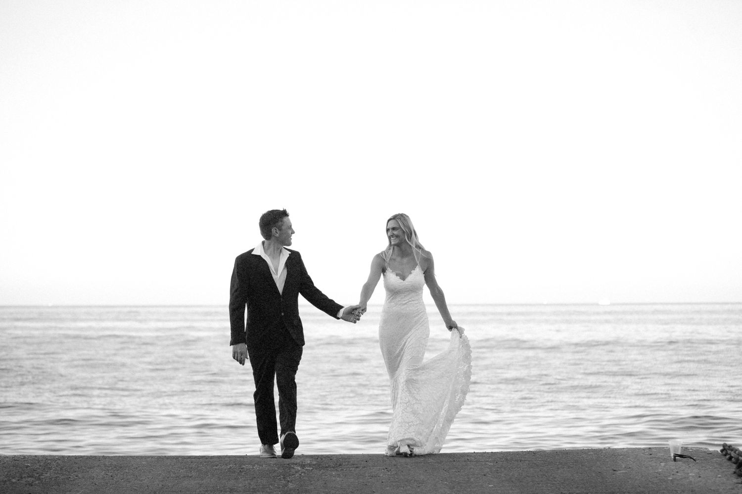 Black and white photo of a bride and groom holding hands walking along a beach shoreline on their wedding day.