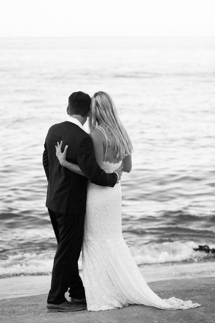Black and white photo of a bride and groom embracing by the ocean, viewed from behind on their wedding day.