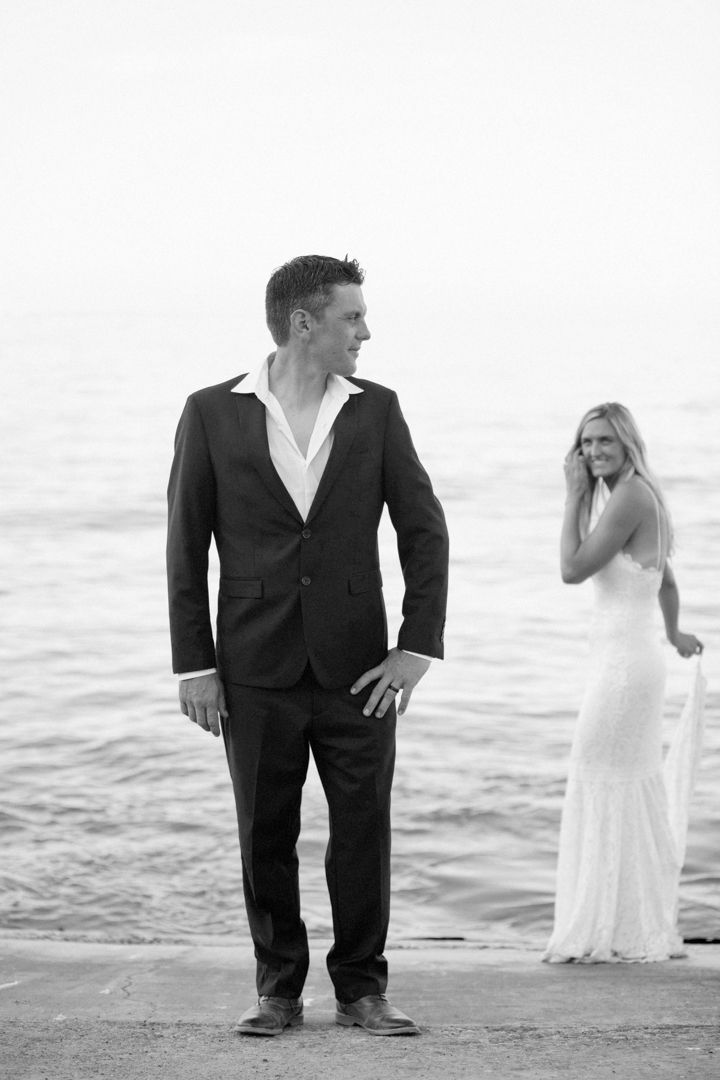 Black and white photo of groom in suit standing on dock with bride in white gown near ocean.