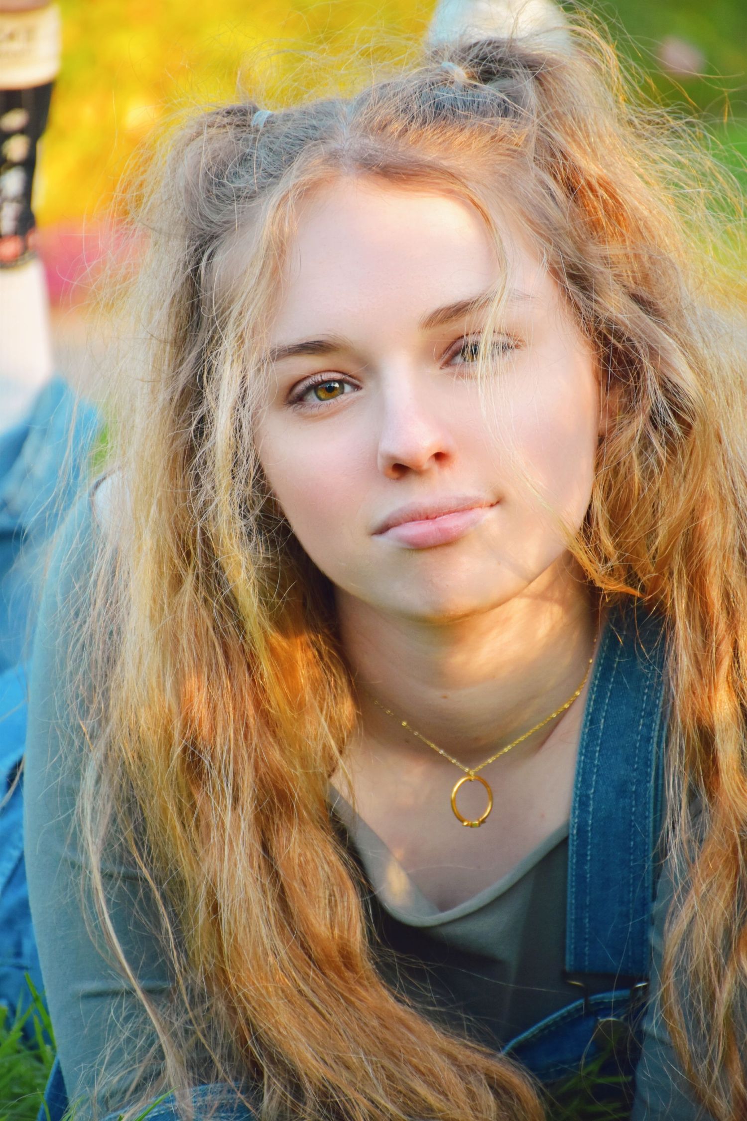 Young woman with long wavy blonde hair wearing denim overalls and gold necklace sitting outdoors in warm sunlight.