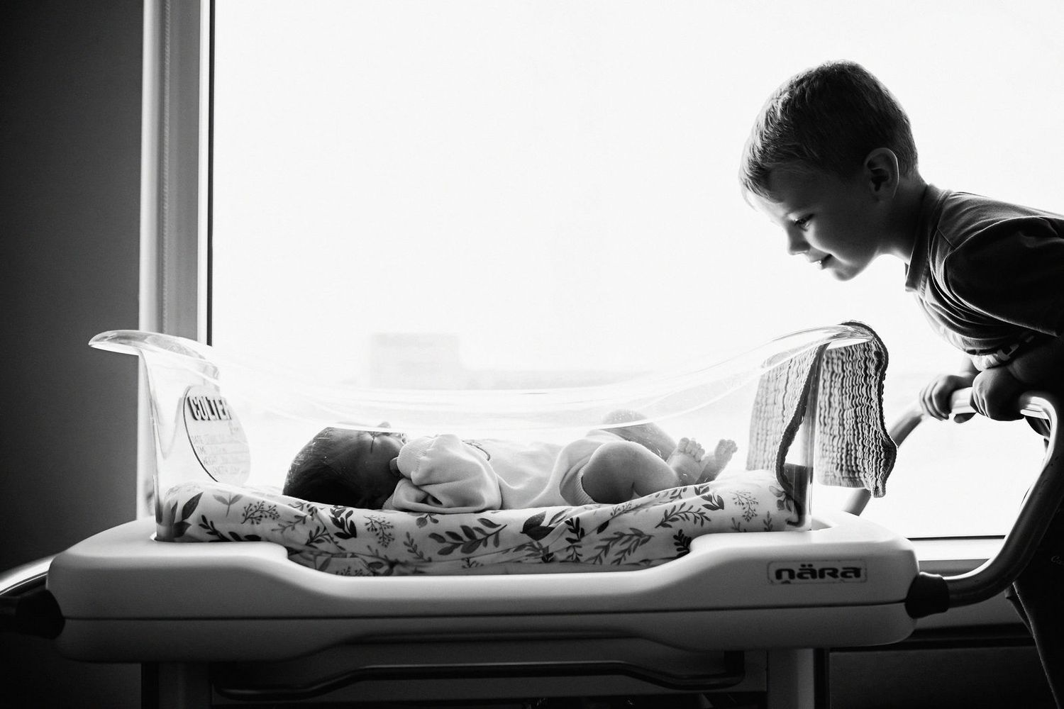 Black and white photo of a young boy leaning over a hospital bassinet to look at a newborn baby.