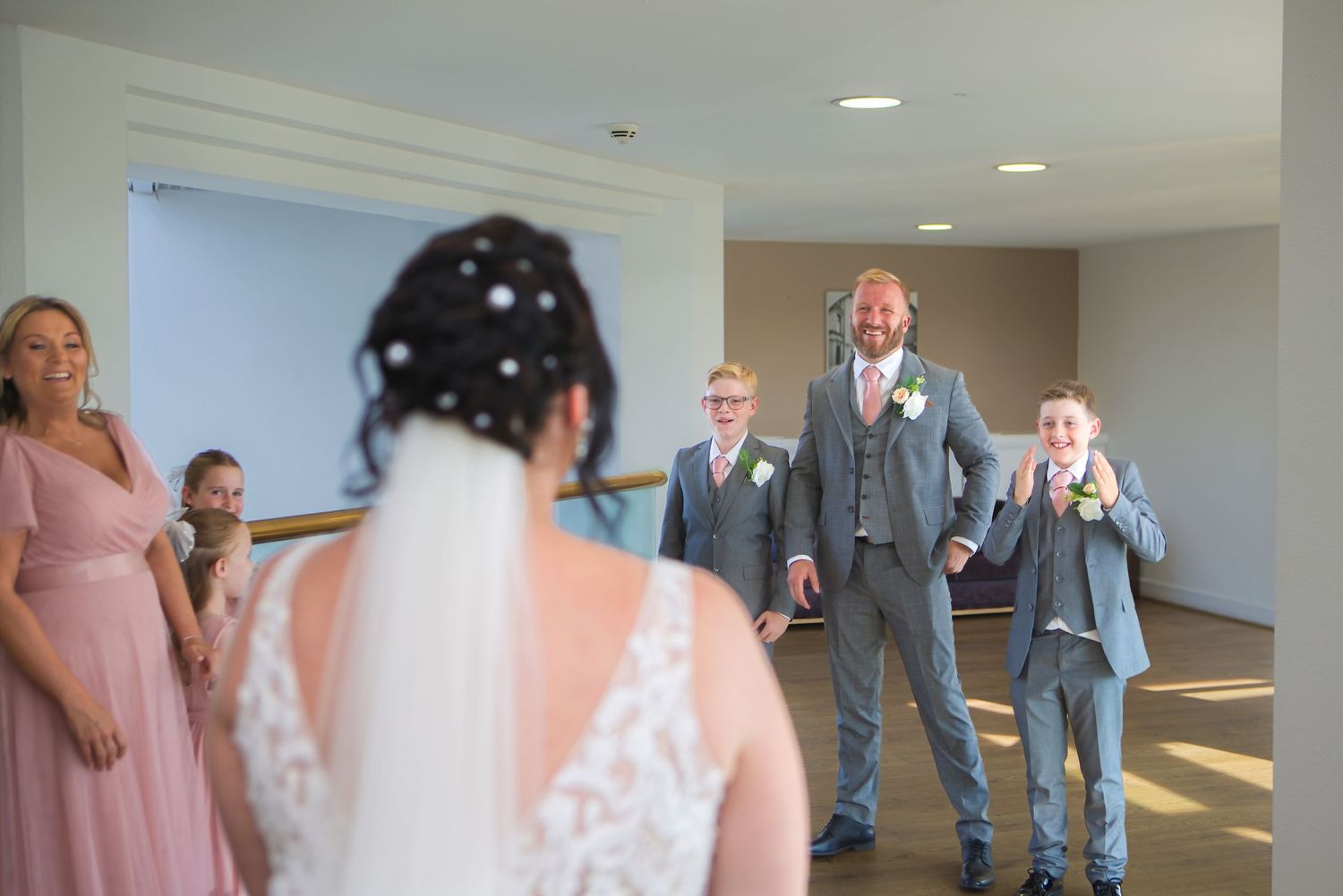 Groom and groomsmen react with joy seeing bride in wedding dress during emotional first look at indoor venue.