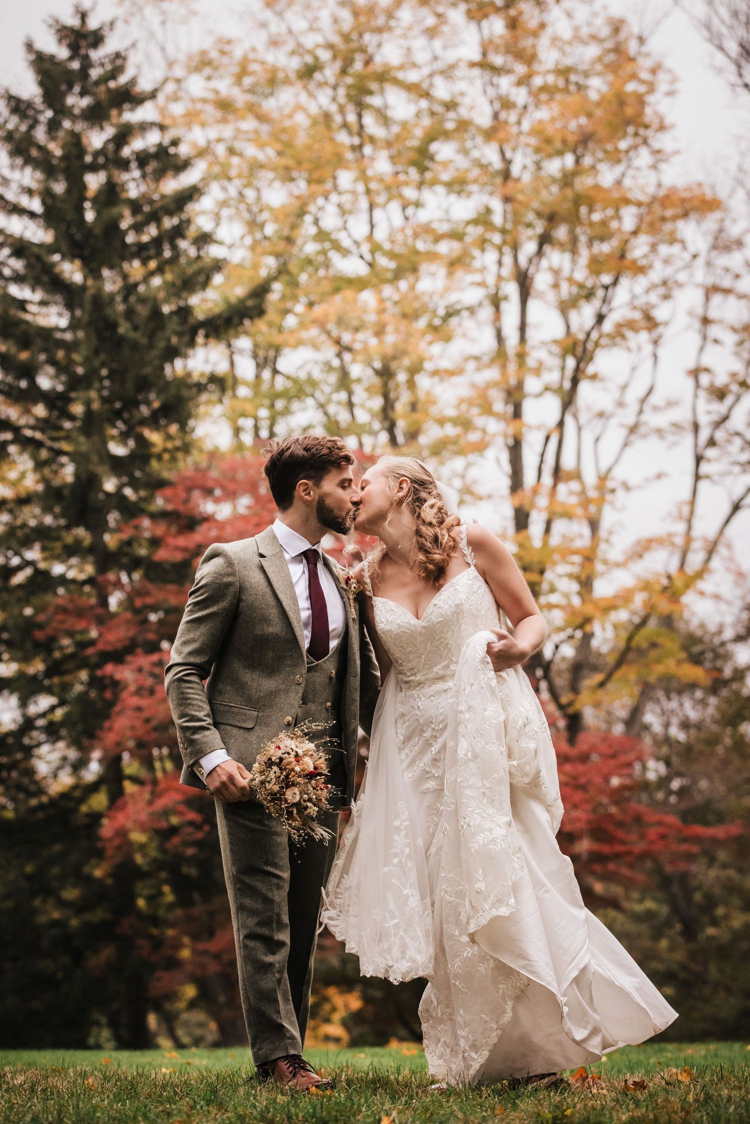 Bride and groom share a kiss outdoors surrounded by vibrant fall foliage in red, orange, and yellow hues.