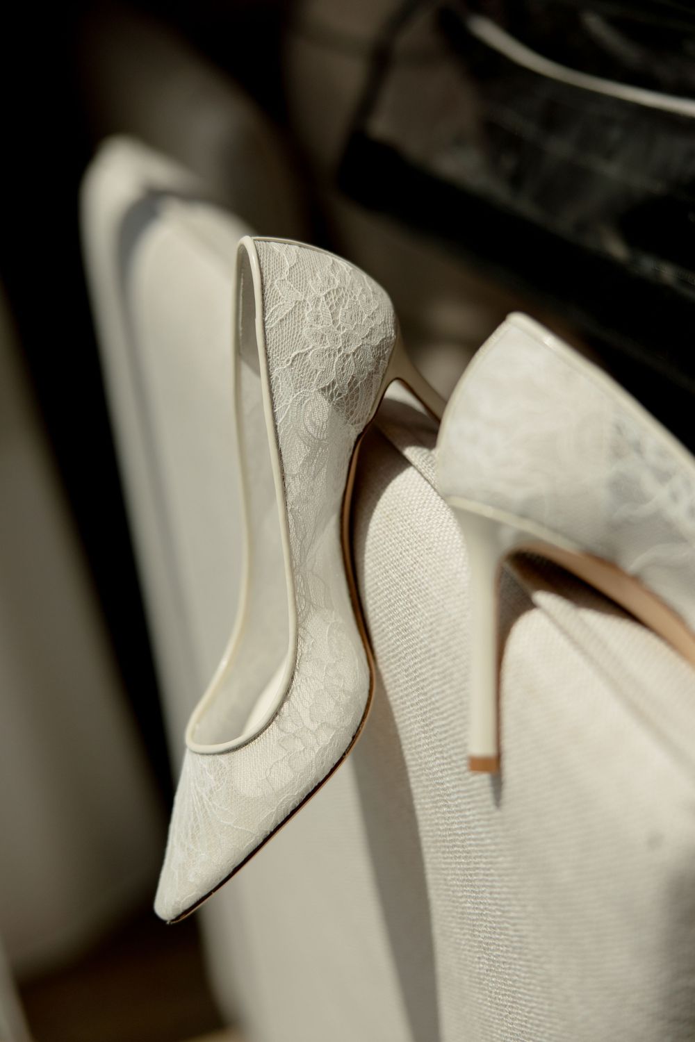 Elegant white lace bridal heels with pointed toe placed on white chair in soft natural light.