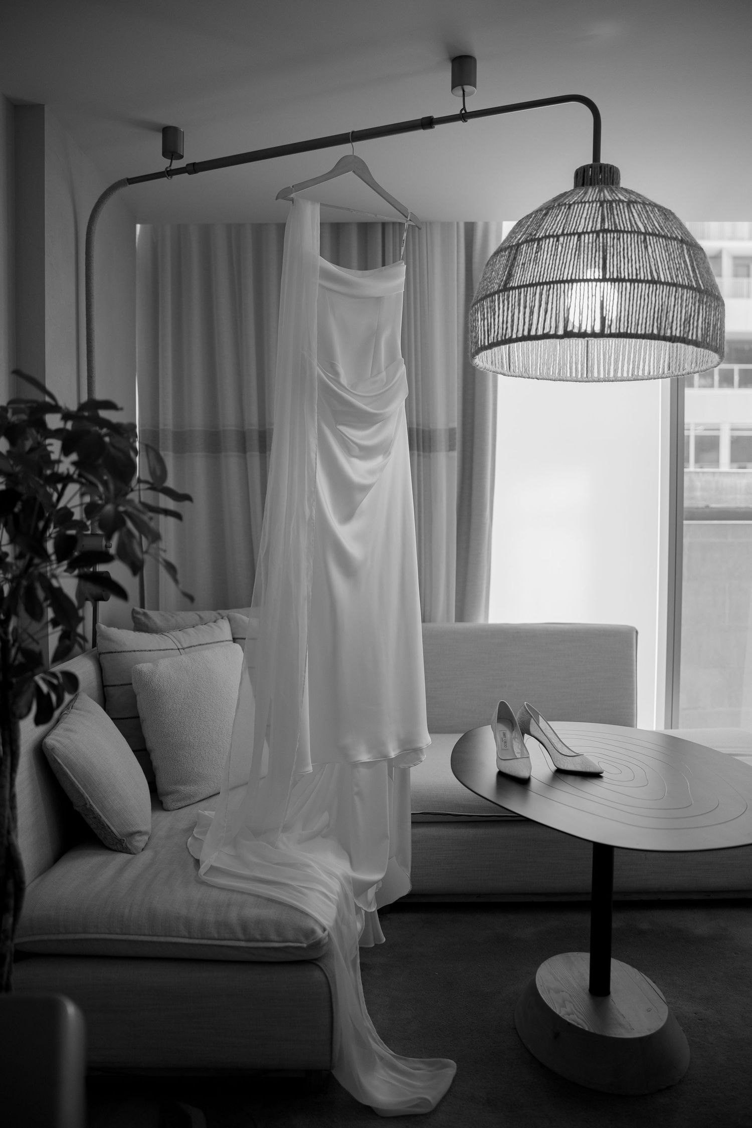 Black and white photo of elegant wedding dress hanging near lamp with bridal heels on table.