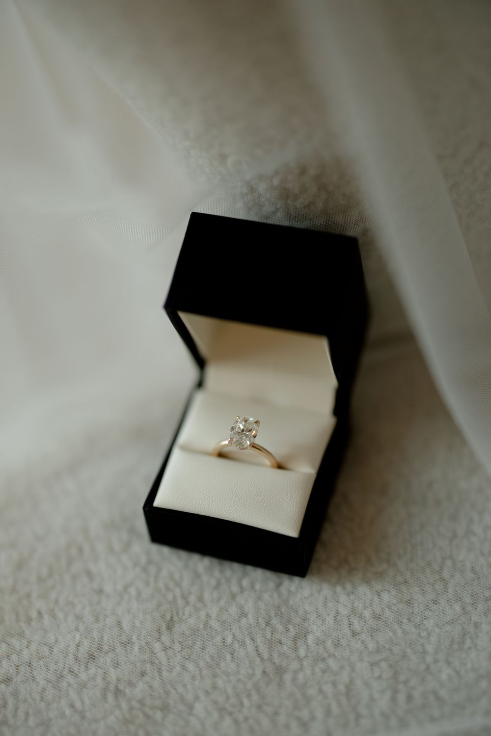 Close-up of diamond solitaire engagement ring displayed in black jewelry box on white fabric.