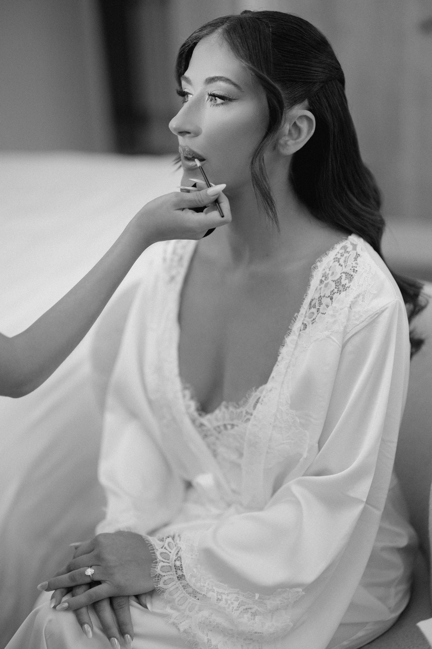 Black and white close-up of makeup artist applying lipstick to bride in white lace bridal robe.
