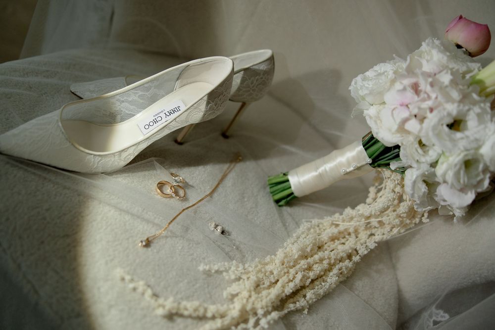 White Jimmy Choo bridal shoe beside pearl necklace, rings, and floral wedding bouquet.