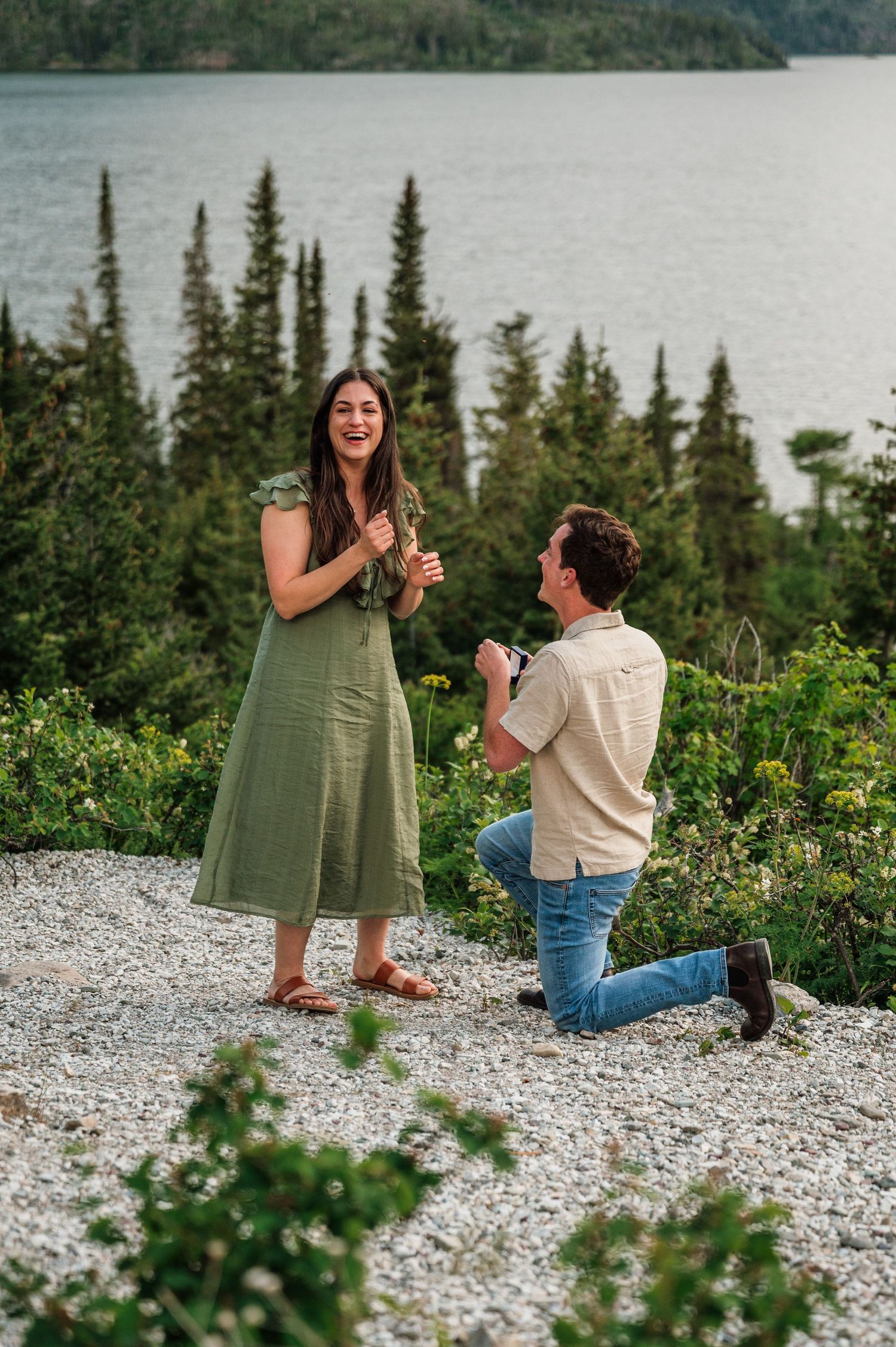 Man proposes on one knee to woman in green dress on rocky overlook with pine trees and lake in background.