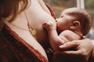 Intimate breastfeeding moment with baby reaching up wearing gold necklace in warm tones.