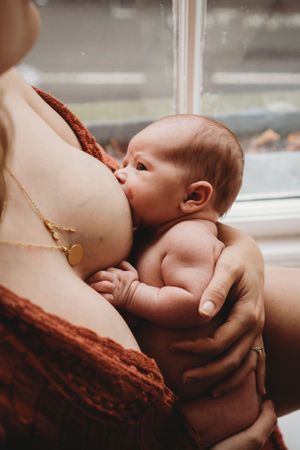 Warm-toned photo of breastfeeding infant wearing gold pendant necklace by window.