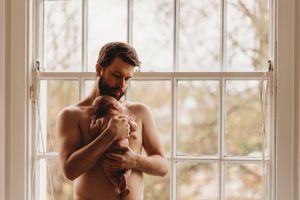 Gentle parenting moment of shirtless caregiver holding infant by bright window.