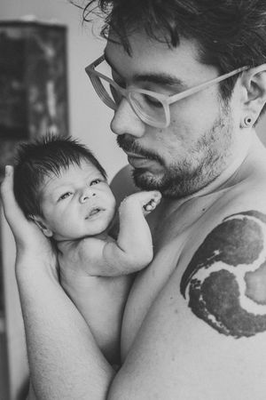 Black and white portrait of a parent holding a newborn baby while showing off an arm tattoo.