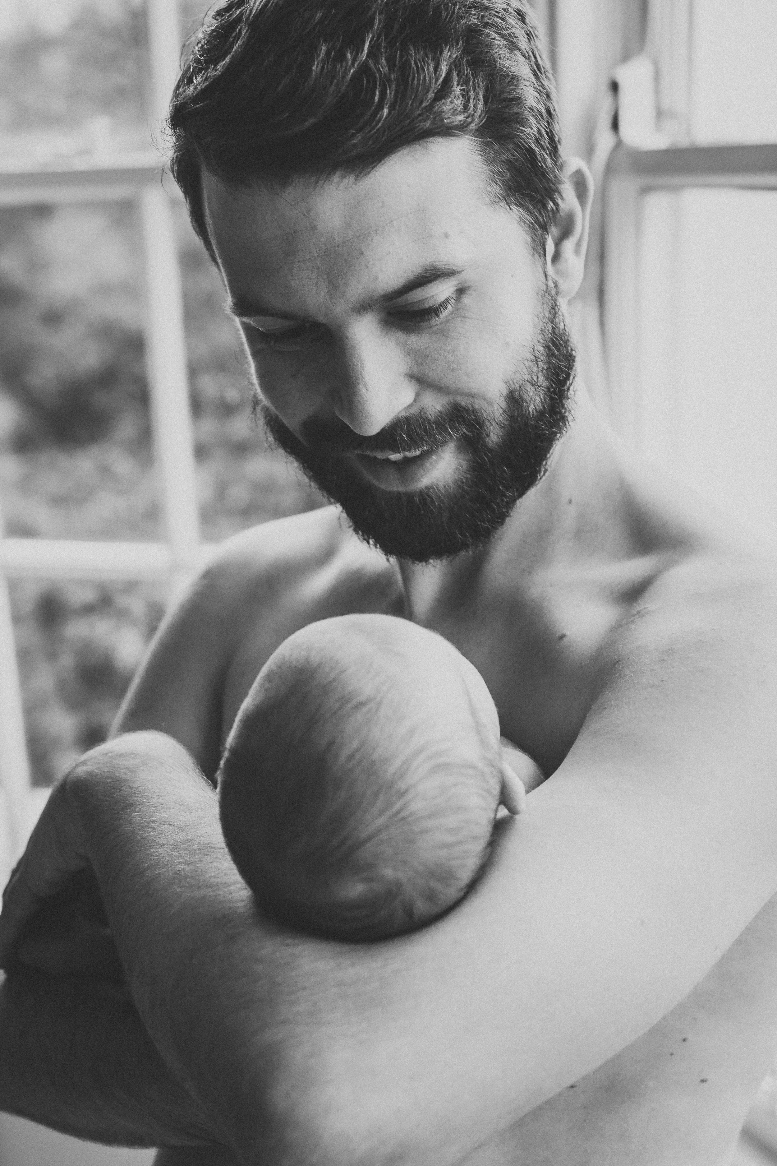 Tender moment of bearded parent holding sleeping infant by window in black and white.