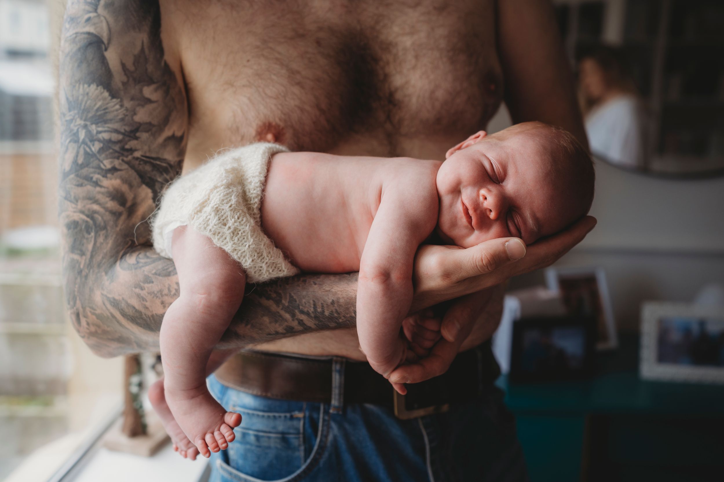 A sleeping newborn baby being cradled by tattooed arms in natural window light.