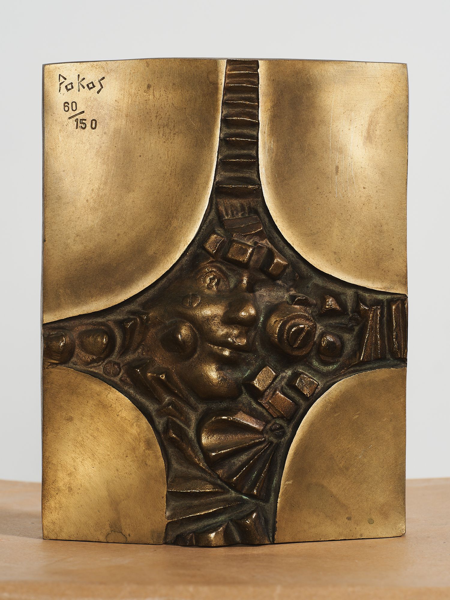 Gold bronze relief plaque signed Pakas 80/150, featuring an abstract face surrounded by geometric patterns.
